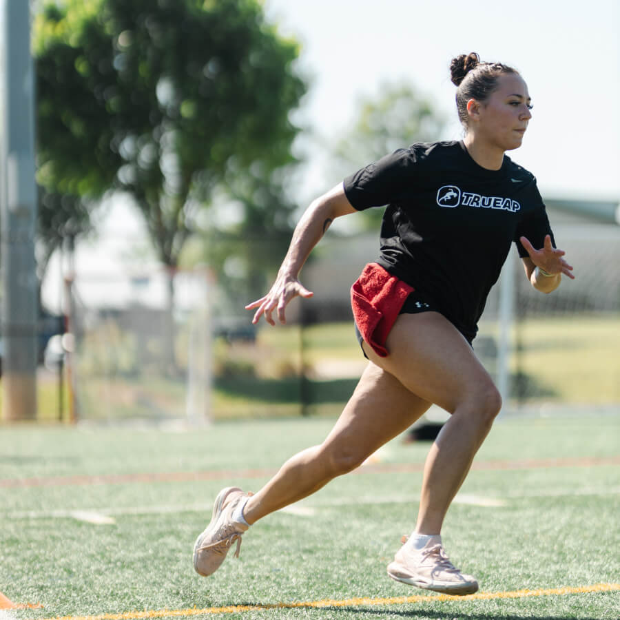 A girl in a black TrueAP shirt runs on a turf field.